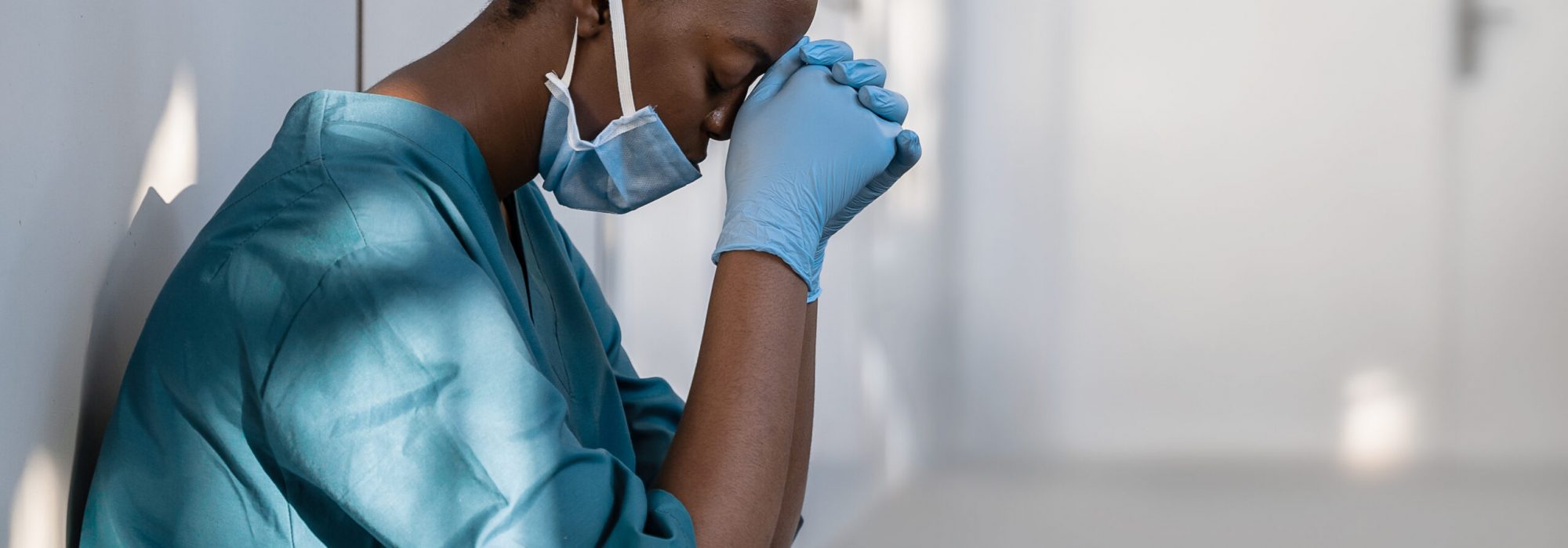 Tired depressed female african scrub nurse wears face mask blue uniform gloves sits on hospital floor. Exhausted sad black doctor feels burnout stress of corona virus frontline protection pray at work